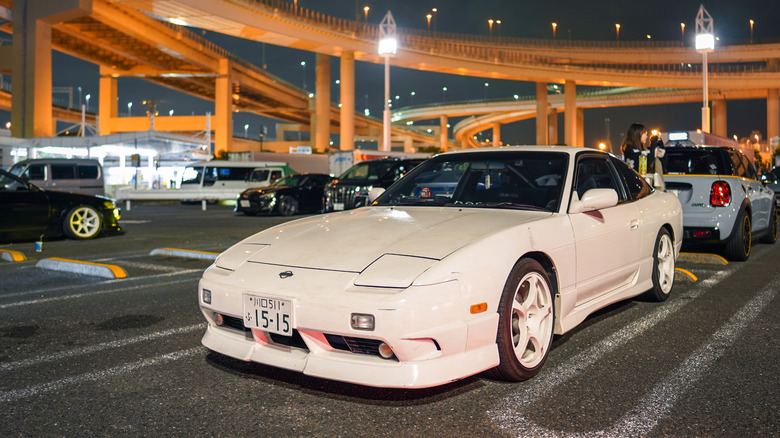 Front 3/4 view of white Nissan 180SX parked at Daikoku Parking Area in Japan.