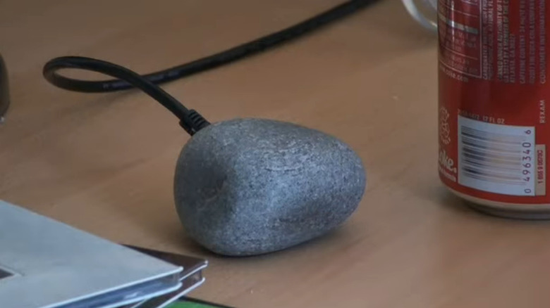 A grey rock plugged into a long black USB cable on a cluttered desk.