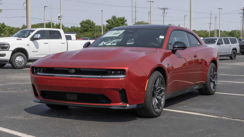 A red Dodge Charger Daytona on a dealership lot, front 3/4 view, heavy duty RAM truck and Jeep Grand Wagoneer in the background