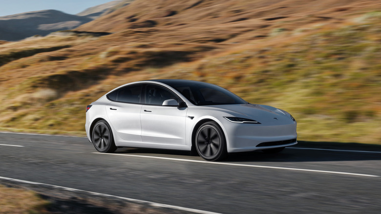 A silver Tesla Model 3 on the road with dry hills behind it