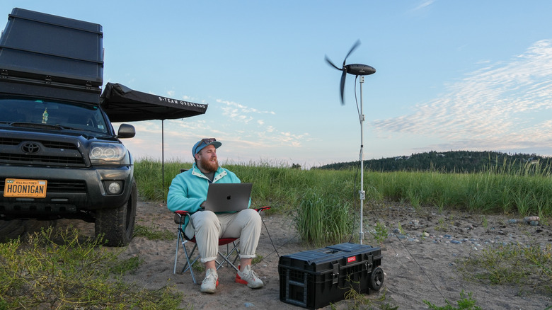 Man sits in a campsite with a laptop and a turbine.