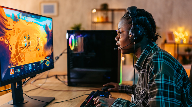 Gamer with headset playing PC game on desktop with PC tower in background, left-side view