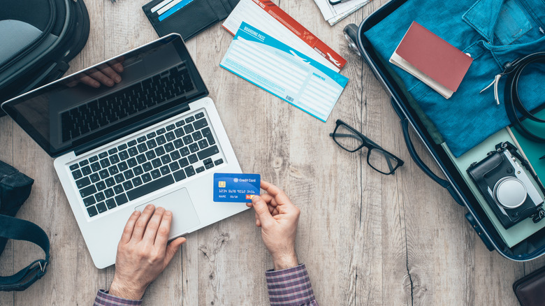 Man preparing for a trip on a laptop while holding a credit card, with a semi-packed suitcase nearby