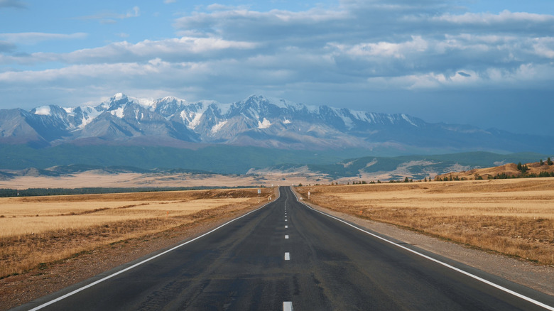 A long open stretch of empty highway running into the desert, with snowy mountains in the background.