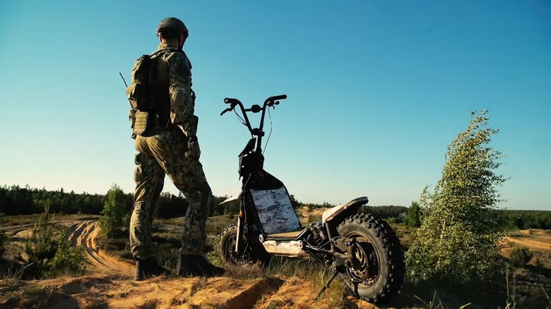 A soldier standing next to a Mosphera e-scooter made by Global Wolf Motors in the battlefield.