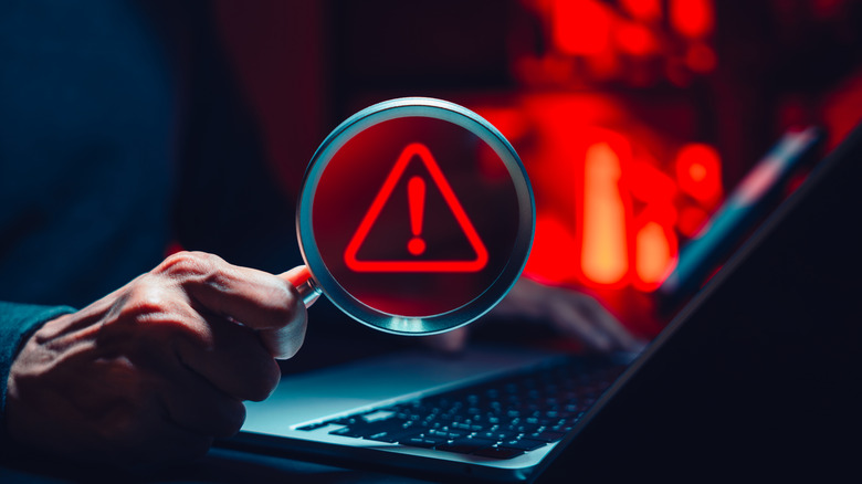 Person holding magnifying glass with digital alert symbol over laptop