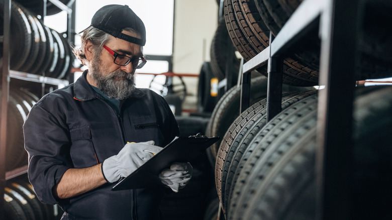 Caucasian man tire technician inspects tire inventory while holding a clipboard in a storage area filled with stacked tires and industrial shelving