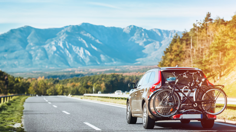 A car driving on a sunny day with bikes mounted to a hitch rack, headed towards woods and mountains on the horizon