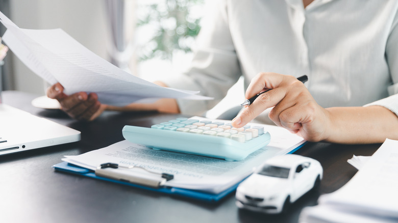 A woman seated at a desk holding papers and using a calculator sitting on top of a clipboard of papers and a small model of a white sedan