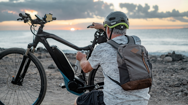 Grey haired man putting water bottle into electric bike, facing ocean sun set