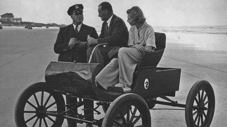 A policeman writes a ticket for a couple riding a 1905 Curved Dash on the beach