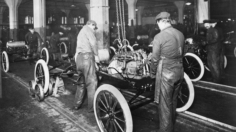 A Model T Ford being assembled, engine lowered from ceiling conveyor belt