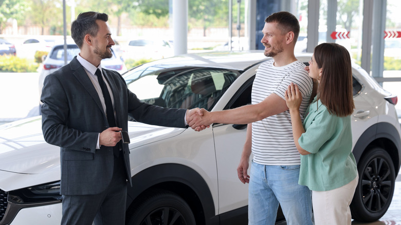 A smile couple shaking hands with a man in a suit in front of a white vehicle inside a car dealership