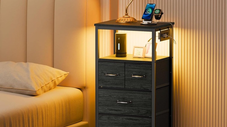 A dresser with a built in outlet and a smart watch and phone plugged in, there are some photos and a lamp on the dresser, the dresser is next to a bed.