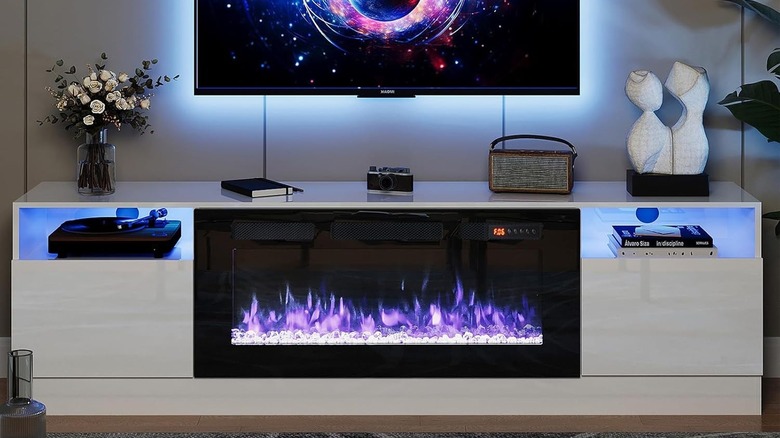 A TV stand with a fireplace inside and a camera, flowers, a radio, a statue, and a notebook on top.