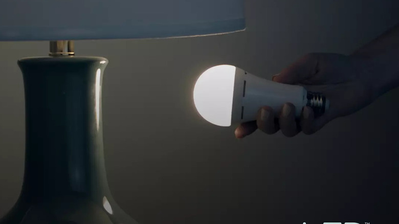 A person holding the GE LED+ Battery Backup Soft White A21 next to a lamp