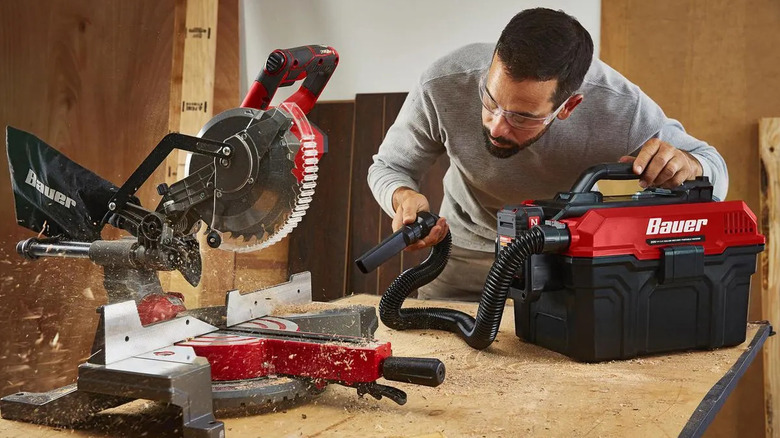A man in safety glasses uses a compact red-and-black Bauer 20V cordless wet/dry vacuum to clean sawdust around a miter saw on a wooden workbench.