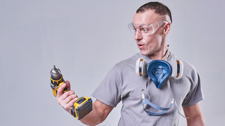 A man wearing safety gear looks at a yellow cordless drill in his hand with a surprised expression.