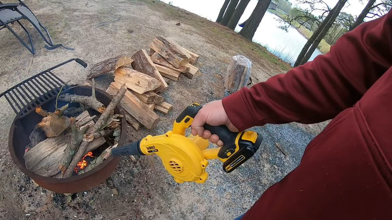 A person uses a DeWalt cordless leaf blower to help ignite a campfire in a fire ring outdoors.