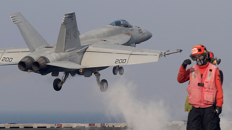 "Red Jersey" crewmember stands in front of jet taking off from flight deck of a an aircraft carrier