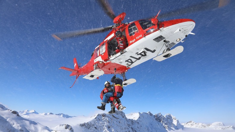 Rega Da Vinci helicopter hovering over mountainous terrain with two people hanging from it