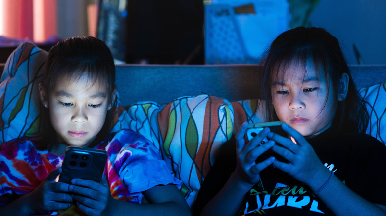 Two children sit on a sofa using smartphones.