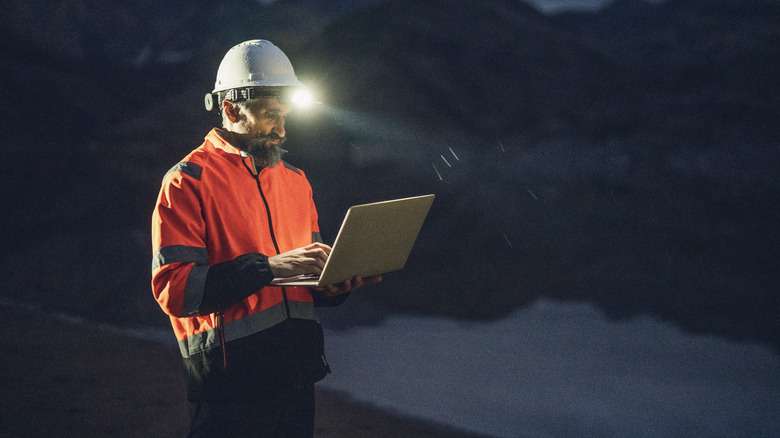 Worker in a hard hat with a light works on a laptop.