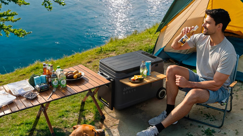 A man drinking a beer while camping next to a lake, sitting next to his Anker Solix smart cooler box.
