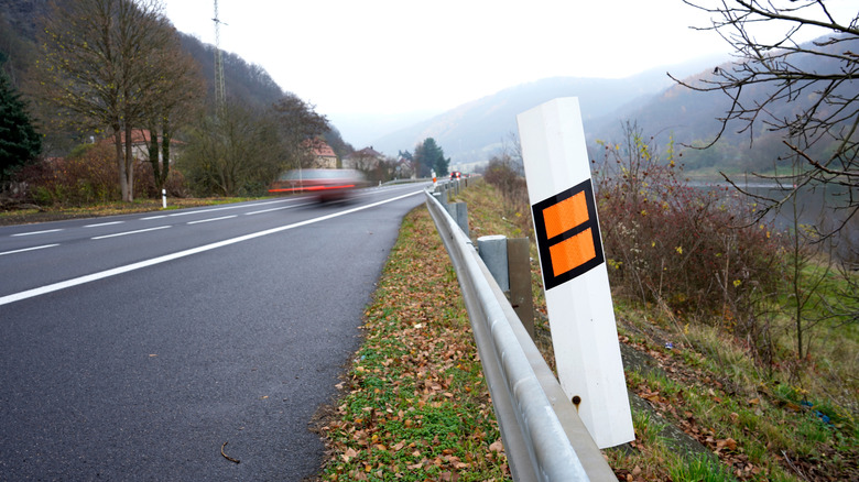 A white reflector post with orange reflective strips on the side of a road, car zooming past