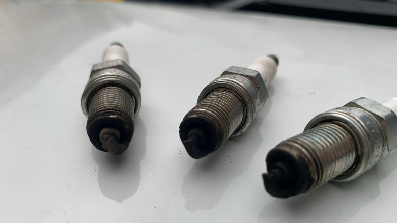Three spark plugs resting on white table, each presenting blackened carbon fouling on tips