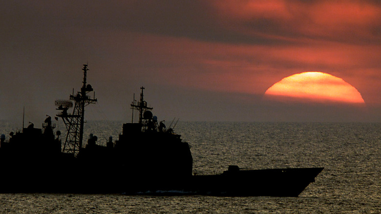 Silhouette side view of a Ticonderoga-class cruiser on the water against a setting sun on the horizon.