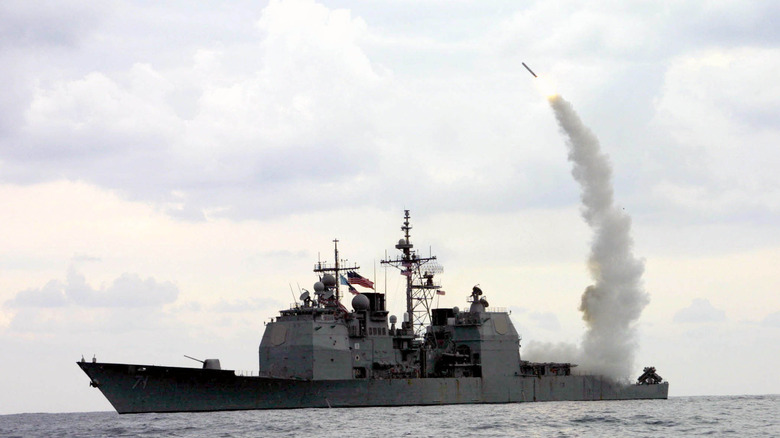 Port side view of a Ticonderoga-class on open water firing a Tomahawk missile against cloudy skies.