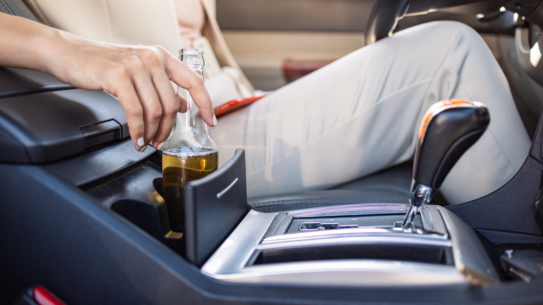 A woman holding bottle of beer while its sitting in the middle console of the car.