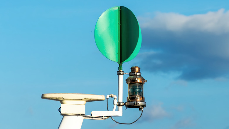 A close-up of a green maritime navigation light and signal lantern mounted on a white boat mast against blue sky and clouds.