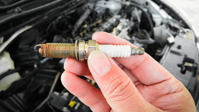 A spark plug being held in a hand with an open car's hood in the background