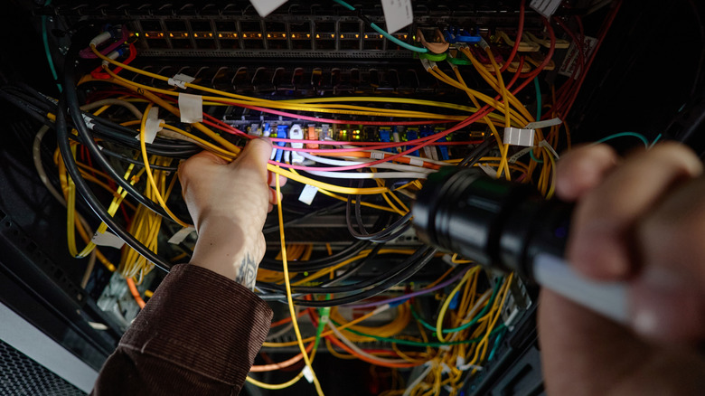 A pair of hands, one holding wires and one shining a flashlight on them on a server rack