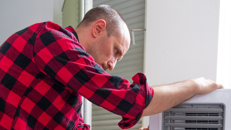 A man in a red and black checkered shirt trying to fix a portable air conditioner