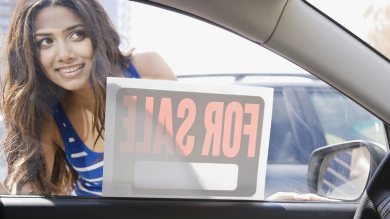 A young brunette woman with brown eyes wearing a blue sleeveless top peers through the side window of a car with a For Sale sign. The picture is taken from inside the car so the lettering on the For Sale sign is shown backward.