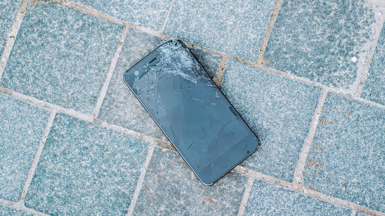 A mobile phone with a cracked screen on a brick pavement.