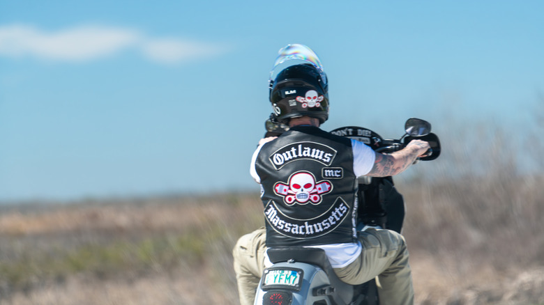 A male driving a Harley Davidson motorcycle wearing a black leather vest with the patch for Outlaws Motorcycle Club for Massachusetts