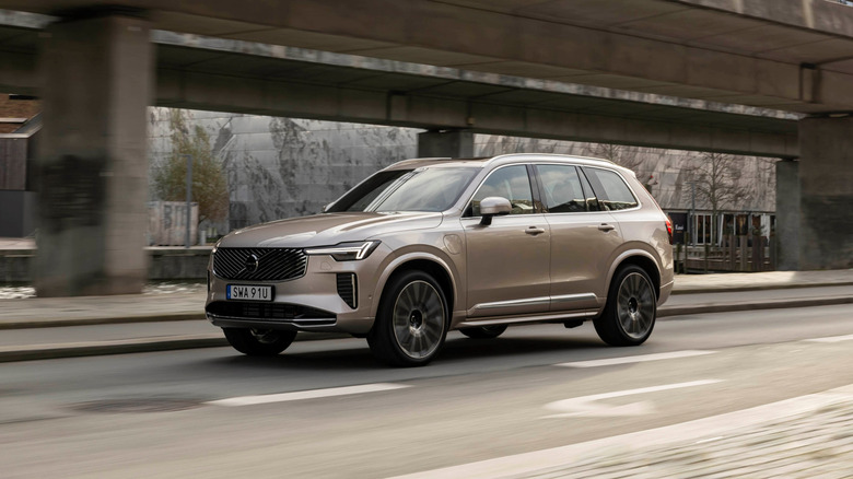 A 2025.5 Volvo XC90 in gold on the move next to a modern bridge, front 3/4 view