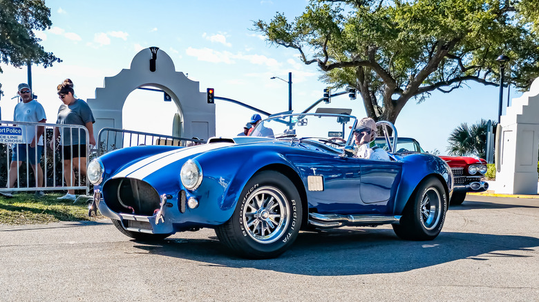 A 60s Shelby Cobra leaving a car show, front 3/4 view, blue with white stripes