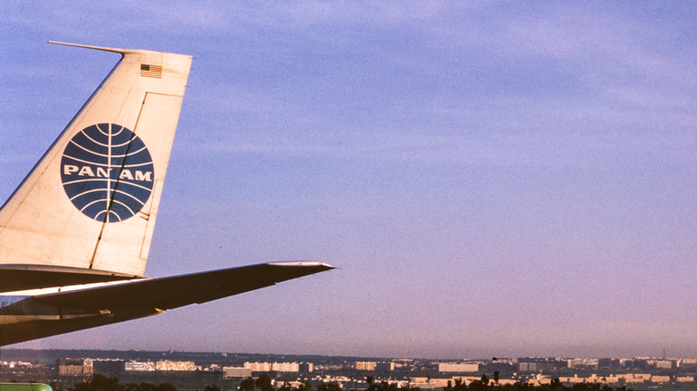 Vintage photo of the tail of a Pan Am plane bearing the Pan Am logo against a blue sky with buildings on the horizon