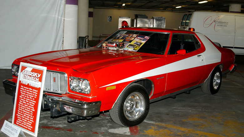 A red Ford Torino with a white stripe is parked in an undercover area with a placard reading 'Starsky & Hutch' against the front grille and paraperhalia on the winscreen.