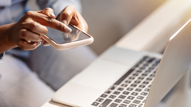 A close-up of a woman using a smartphone and laptop at the office table.