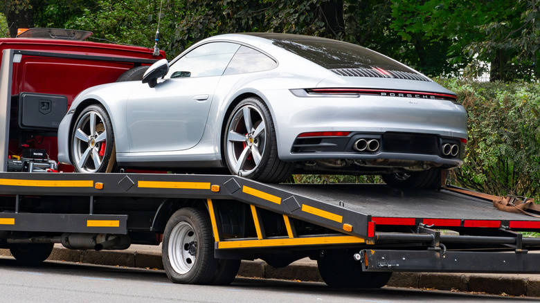 A silver Porsche 911 loaded on a black and red flatbed truck with trees in the background.