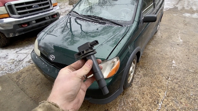 man holds up a coil pack in front of a green car