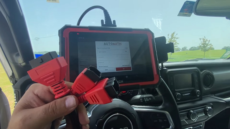 Driver holding a Cummins bypass harness with a tablet showing the AutoAuth login screen in the background