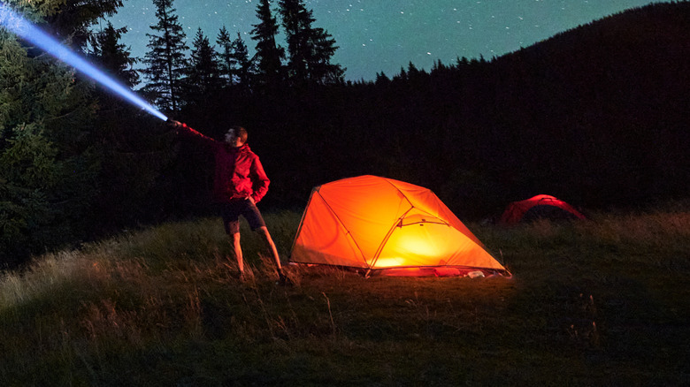 A man on a grass field using a flashlight and pointing the beam towards the night sky, as he stands next to camping tent