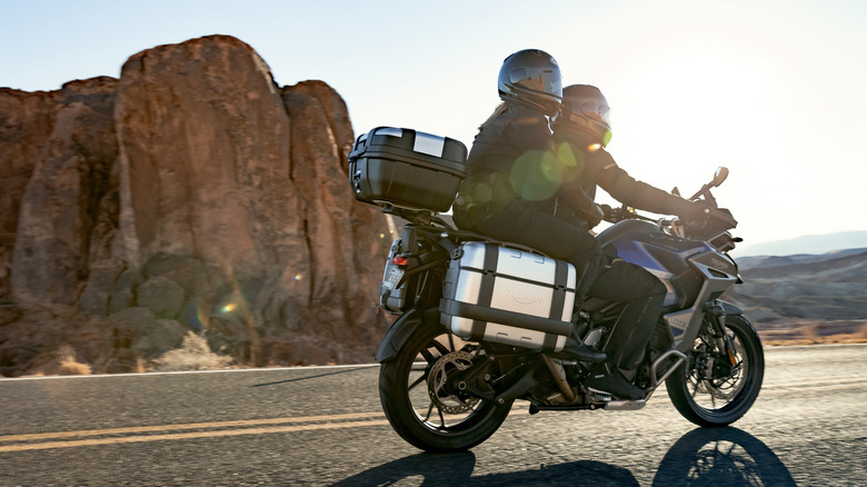 A touring motorcycle rider and passenger on a highway with a rocky backdrop.